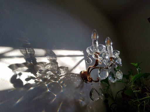 The photo shows a chandelier with crystal pendants casting beautiful reflections and shadows on the wall. The sunlight is shining through a window, highlighting the details of the chandelier and its ornate design, with light sparkling off the crystals. The warm light creates intricate patterns on the surrounding wall, adding a serene and artistic atmosphere.