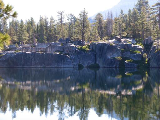 Large granite stone walls form the edge and reflect in a calm lake, with trees and shrubs.