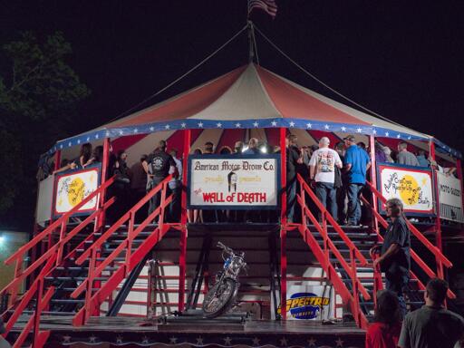 A large elevated tent with a group of people down inside watching the wall of death show where motorcycles ride along the inner walls doing stunts