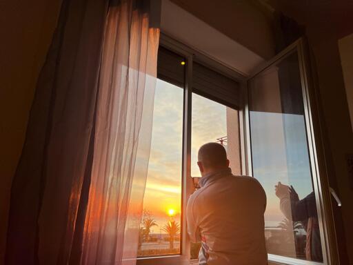 A person (me) taking a photo of a beautiful sunrise through a window with sheer curtains, capturing the moment while a reflection of their hand holding the camera is visible on the glass.