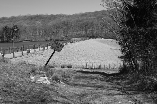 A black and white photo of the back of the dam at South Mountain Reservation. A no trespassing sign is in the foreground. The dam is just visible on the left of the image, and trees coming into bloom are in the background.