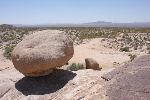 In the foreground, a sandstone-looking boulder balances precariously atop of a rock formation. Behind it the Mojave desert stretches out to low mountains in the distance.