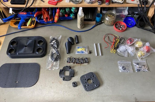 Parts for the Open Source Arcade Stick spread out on my workbench. The case parts are 3d printed, there are some buttons and microswitches, a PCB for auto fire and various small bits and pieces.