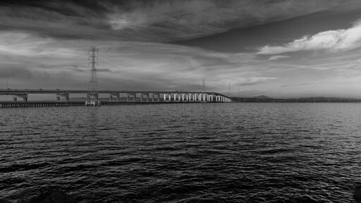 A relatively unremarkable bridge crossing a large body of water. Power pylons run adjacent to the bridge.