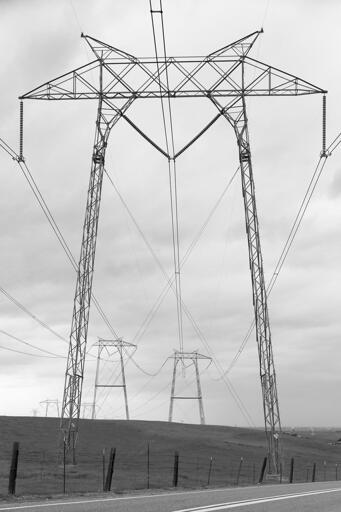 A high-voltage power transmission line "portal" style pylon in a field along a road. Other pylons are visible in the background.