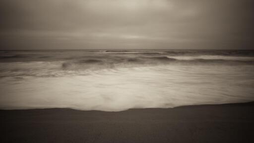 Small waves breaking in the Pacific Ocean, viewed from a beach, on an overcast day.