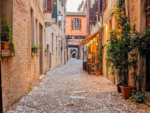 A picturesque cobblestone alley in Ferrara, Italy, bathed in the warm glow of evening lights. The rustic brick walls, adorned with green shutters and potted plants, frame a charming "osteria" with golden-hued awnings and softly lit tables spilling onto the street. Overhead, delicate string lights twinkle, casting a cozy ambiance as the narrow passageway leads into the depths of the historic town. The scene exudes timeless Italian charm, inviting quiet strolls and lingering conversations over a glass of wine.