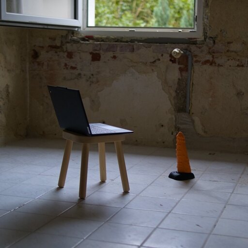An empty room with torn walls and dirty tiles on the floor. In the center is a Lenovo Thinkpad resting on a small stool. A large dildo is placed on the floor where a chair would be if this were a normal desk.