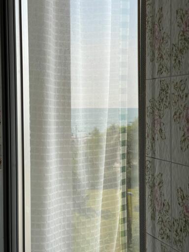 A softly lit window with a sheer white curtain, partially revealing a blurred view of the sea and greenery outside. The reflection on the glass creates a layered perspective, blending the interior and the outside world.