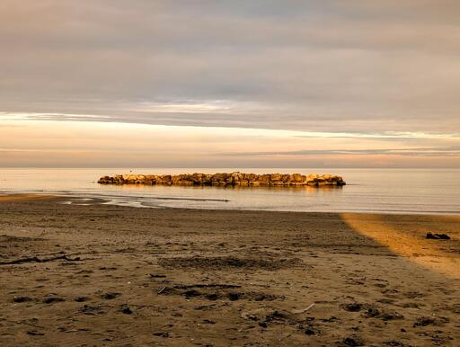 A quiet winter shoreline bathed in the golden light of the setting sun. The sea is calm, its surface reflecting soft hues of amber and rose, while the breakwater glows with the warmth of the fading day. A patch of sunlight stretches across the sand, a fleeting promise that winter is loosening its grip - pring is not so far away.